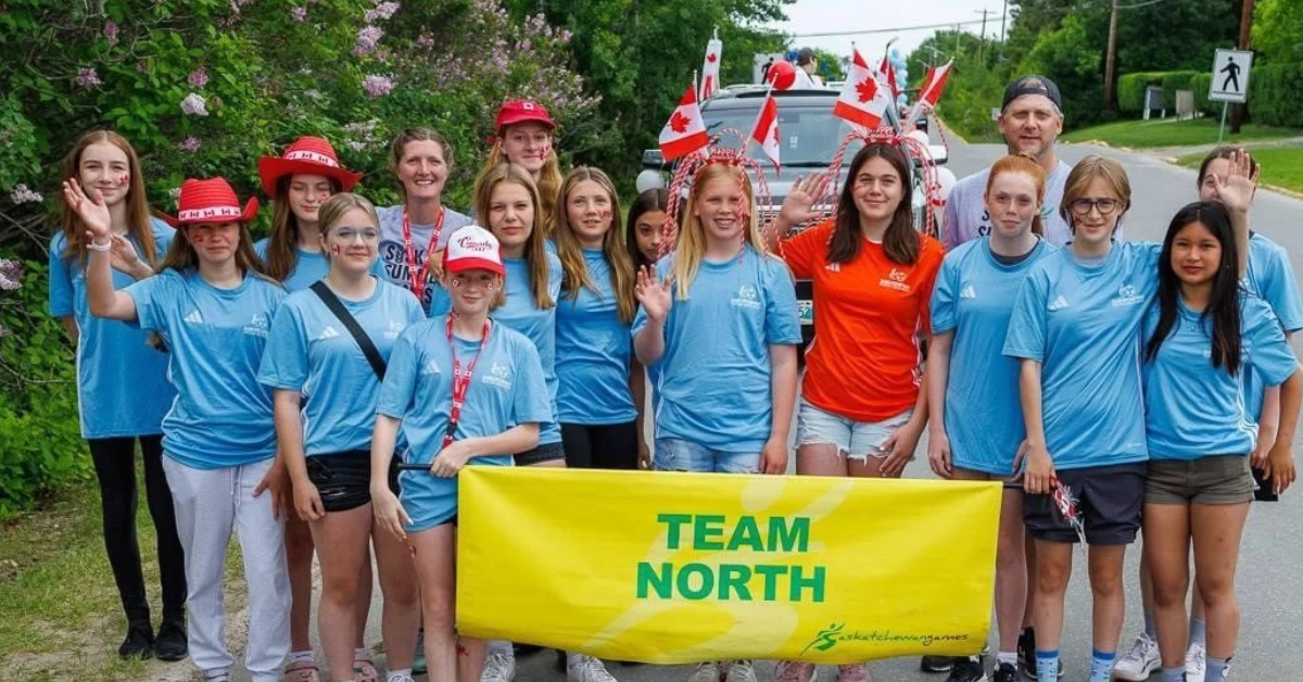 Team North members standing as a group all in light blue shirts, holding a large bright yellow sign that reads Team North in green. Various members are smiling while wearing Canada themed hats and waving Canadian flags.